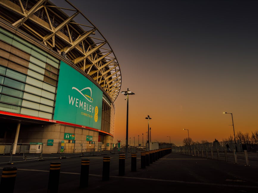 A wide view of Wembley Stadium's football pitch during daytime, featuring vibrant green turf with clearly marked white lines. Surrounding the pitch are rows of red stadium seats, which are clean and w