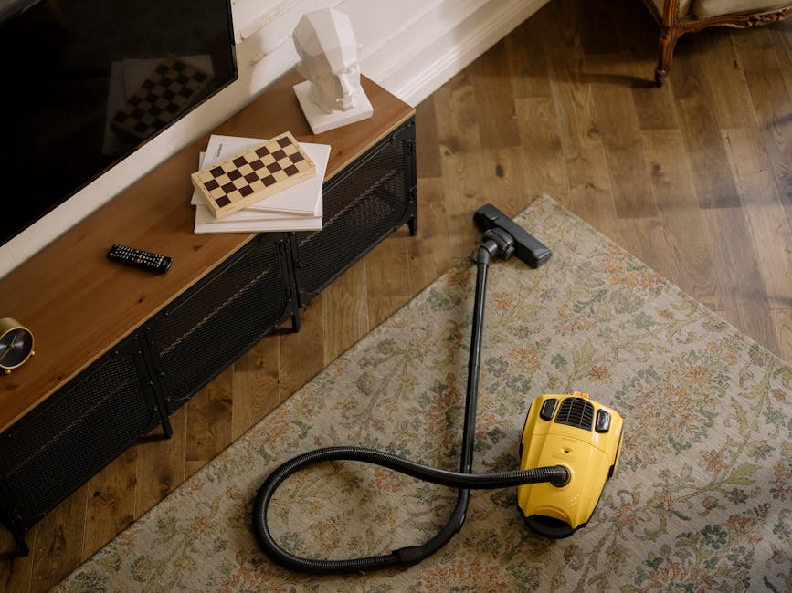 Close-up of a modern vacuum cleaner with a transparent dust container filled with debris, positioned on a pink carpet in a domestic setting. The vacuum has a flexible, patterned hose connected to a fl