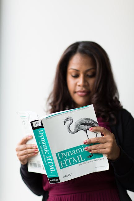 A woman with long dark hair, dressed in a black blazer and maroon top, is holding a book titled 'Dynamic HTML' with a picture of a flamingo on the cover. She is standing indoors against a plain white